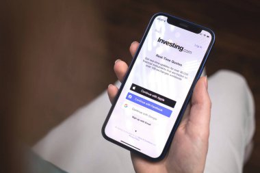 Kharkov, Ukrayna - 10 Ocak 2022: Kadın Investing.com uygulaması kullanıyor. Apple iPhone 12 ile el, logolu cep telefonu