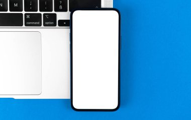 Üst görünüm modeli. Beyaz ekranlı akıllı telefon, dizüstü bilgisayarlı düz mavi arka plan. Asgari çalışma alanı, modern dizüstü bilgisayar, kopyalama alanı