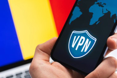 Romanya 'da VPN. Güvenli ve güvenli internet konsepti. Mahremiyet. Cep telefonu ve VPN uygulaması olan bir el. Arka plandaki fotoğrafta bayrak ve dizüstü bilgisayar.