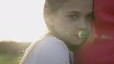 Mavi gökyüzü arka planında annesine mercekle sarılan küçük tatlı bir kızın portresi. Kız gülümsüyor, yaz günü annesiyle dışarıda eğleniyor.