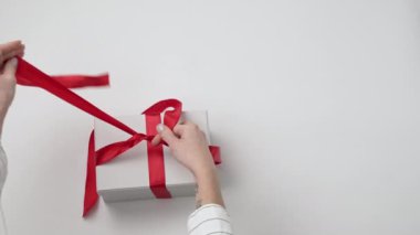 Kadın elleri hediyeyi süslüyor, hediye kutusuna beyaz arka planda bir fiyonk bağlıyor. Kadın eli hediye paketleri. Noel, yeni yıl, doğum günü, tatil, yıldönümü konsepti. Üst manzara, düz uzanma.
