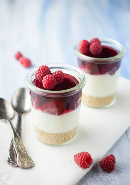 Raspberry cream cheese dessert with a cookie bottom served in a glass on blue background.