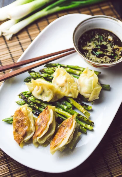 Domuz etiyle dolu bir tabak dolusu gevrek gyoza köftesi ve koyu bir tabakta çekilmiş yeşil kuşkonmazla servis edilmiş.