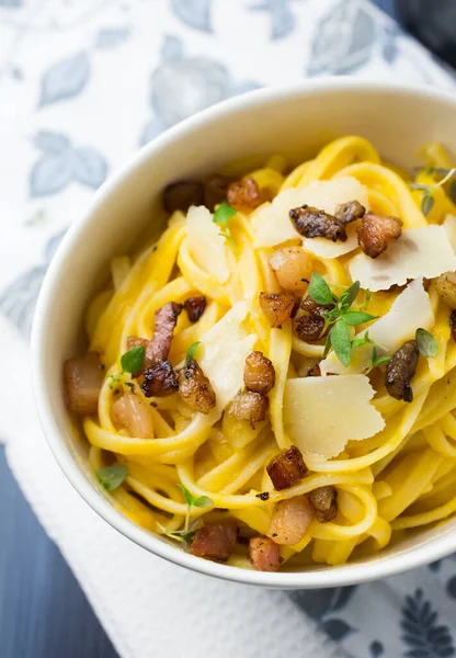 Beyaz kâse spagetti carbonara ve üzerinde mavi gri arka planda parmesan peyniri olan pastırma.