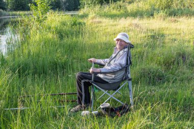 Yaşlı bir balıkçı, güneşli bir yaz gününde sakin bir nehrin kıyısındaki balıkçı zincirinde oltalarla oturur.