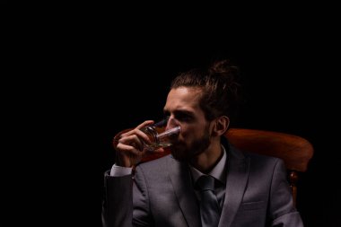 Akıllı gri takım elbiseli genç bir işadamı kasvetli ve atmosferik ışıklandırması olan tahta bir koltukta oturuyor ve tek bir kadeh viski içiyor.