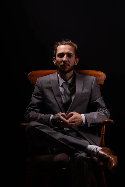 A young businessman in a smart gray suit sitting in a wooden arm chair with moody and atmospheric lighting