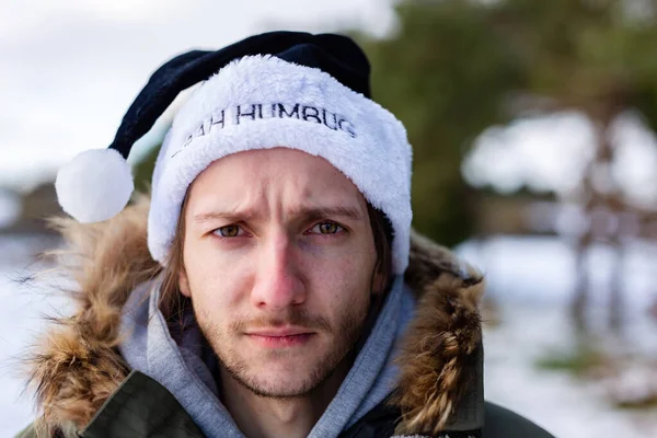 A grumpy young man wearing a black santa hat with the words Bah Humbug written on it