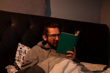 Genç bir adam yatağa uzanıp uyumadan önce kitap okuyor, karanlık bir konseptten sonra.