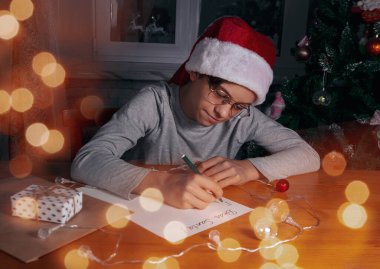Noel Baba gibi kırmızı şapkalı bir çocuk Noel ağacının yanındaki tahta masaya mektup yazıyor..