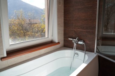 Pencere kenarında su olan saf beyaz bir küvet. Büyük pencereli banyo. Dağ manzarası