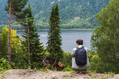 Köpekli bir adam gölün kenarında oturuyor ve doğanın tadını çıkarıyor.
