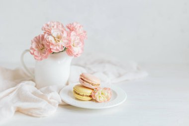 İki lezzetli Fransız makaronu ve beyaz arka planda pembe karanfil çiçekli bir kavanoz. Pastel renkler. Bahar arkaplanı.