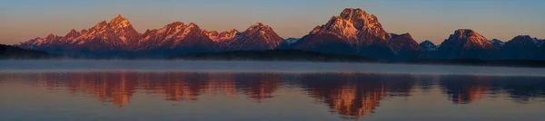 The mountain range of Grand Teton National Park is photographed during a sunrise with the peaks\' reflections in Jackson Lake on a cloudless summer day.
