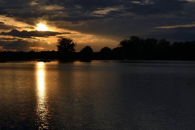Gölde yağmur bulutları ve ağaç siluetleriyle yaz günbatımı