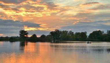Bir çayırdaki gölün üzerinde gün batımı bulutlu gökyüzünün sularında bir yansımayla, bir balıkçı.