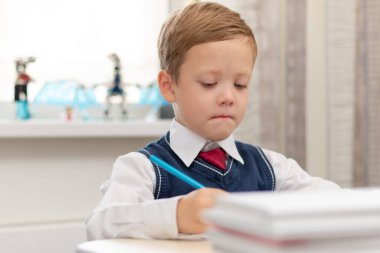 Birinci sınıf üniformalı tatlı bir çocuk evde elinde kalemle masada otururken ev ödevi yapıyor. Uzaktan eğitim. Seçici odaklanma. Yakın plan. Portre