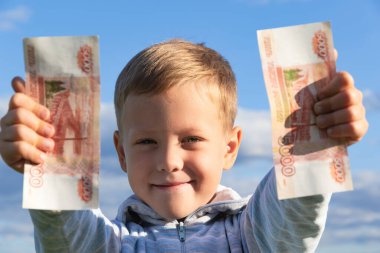 Süveter giyen tatmin olmuş bir anaokulu çocuğu, bulutlu mavi gökyüzünün arka planına karşı doğada kağıt ruble taşır. Yakın plan. Portre