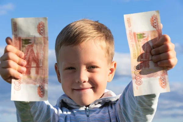 Süveter giyen tatmin olmuş bir anaokulu çocuğu, bulutlu mavi gökyüzünün arka planına karşı doğada kağıt ruble taşır. Yakın plan. Portre