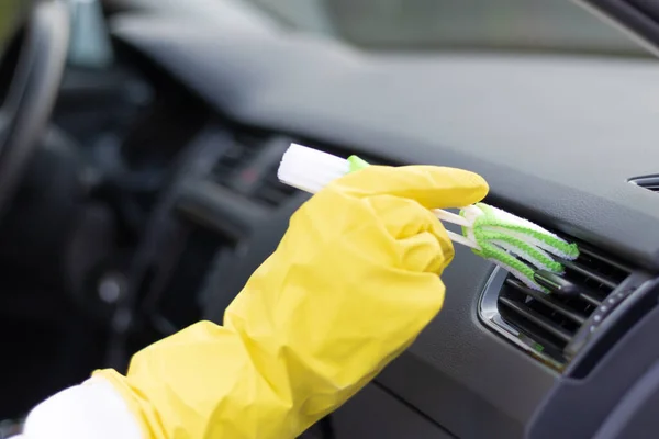 A hand in yellow rubber gloves wipes the dashboard of a car from dust with a special brush on a bright autumn day. Selective focus. Close-up