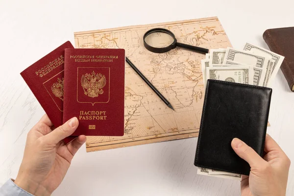 女性的手拿着两张护照和一个装有钱的黑色皮夹 在一张白色的木制桌子上 放着一张世界地图 一个放大镜 一支铅笔 一本日记和一个装有钱的黑色皮夹 护照俄罗斯联邦 — 图库照片