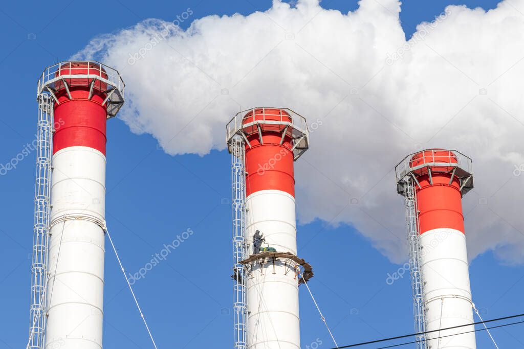 red-white chimneys of the boiler room, equipped with a traffic light. industrial climbers carry out routine repairs. white smoke on a blue sky background