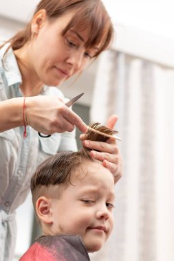 Açık mavi elbiseli, elinde tarak ve makasla güzel bir anne oğlunun saçını evde kesiyor. seçici odak