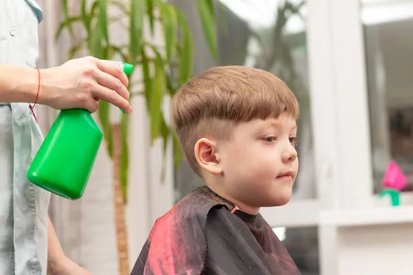 美容室の女性の手は 窓や緑の植物を背景に髪を切る前に 子供の頭の上に緑色のスプレーから水を噴霧します 選択的焦点 — ストック写真