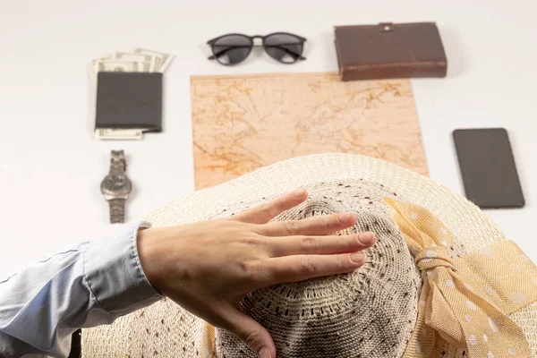 female hand holds a straw hat. on a white wooden table lies a world map, a black wallet with money, a diary, a watch, a smartphone and sunglasses. selective focus