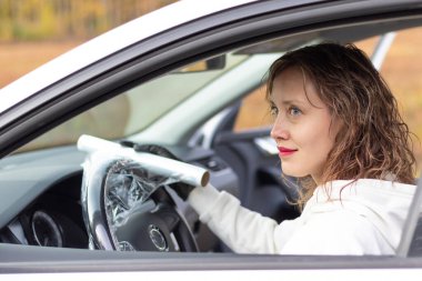 Beyaz ceketli ve siyah eldivenli güzel genç bir kadın sürücü, parlak bir sonbahar gününde servise gitmeden önce bir arabanın direksiyonunu koruyucu bir filmle sarar. Seçici odaklanma. Portre