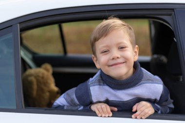 Yedi yaşında tatlı bir çocuk, güneşli bir sonbahar gününde beyaz bir arabanın camından sarkan sarı yapraklara yaslanıyor. Seçici odaklanma. Portre