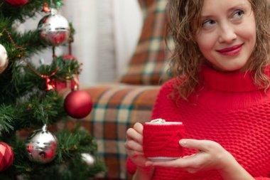 Yılbaşı arifesinde yeni yıl ağacının yanında elinde bir kupa kakao ve marşmelovla kırmızı kazaklı güzel bir kadın. Seçici odaklanma. Portre