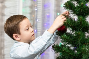 Yedi yaşında mavi gömlekli bir çocuk, Noel ağacını Noel arifesinde evde çelenklerle süslüyor. Seçici odaklanma. Portre