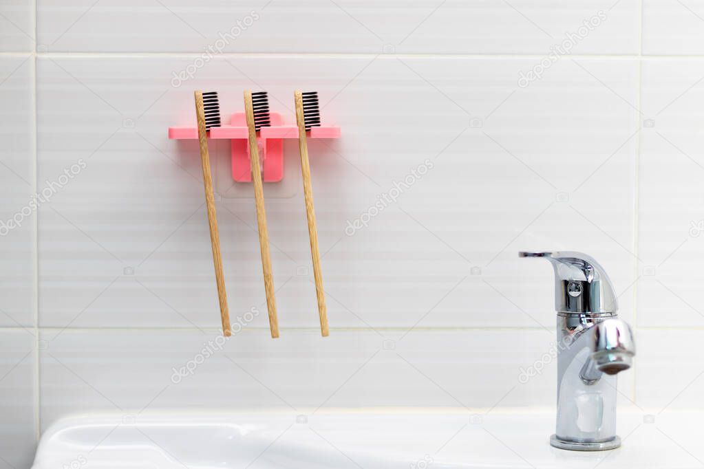 Three natural bamboo toothbrushes with black bristles in a holder against a tile background on the bathroom wall above the washbasin. Selective focus. Close-up