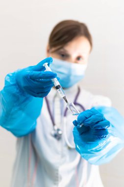 Tıbbi maske takmış genç bir kadın doktor ve elinde şırıngayla eldivenler ve koronavirüs salgını sırasında hastanede steteskop. Seçici odaklanma. Portre