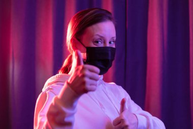 Siyah tıbbi maskeli ve mavi menekşe arka planda beyaz kapüşonlu süveter giyen genç ve güzel bir kadın bir koronavirüs salgını sırasında el hareketlerini gösteriyor. Seçici odaklanma. Portre