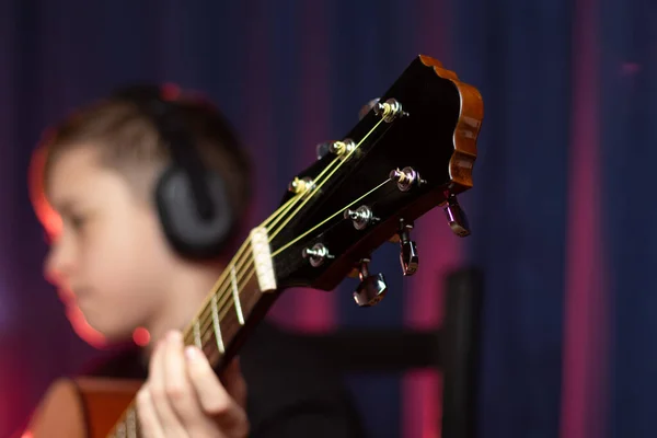 一名13岁的少年身穿黑色衬衫 头戴耳机 在音乐演播室里带着一把吉他 在紫色背景下记录了一支带红色照明的曲调 有选择的重点 肖像画 — 图库照片
