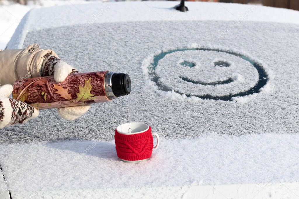 Women's hands in mittens pour boiling water into a mug from a thermos on a snowy car trunk on a sunny winter day. Smiley drawing. Selective focus. Close-up