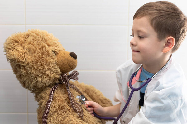A cute boy of 7 years old in a medical gown with a stethoscope checks the health of a toy bear. Doctor and hospital game. Selective focus. Portrait