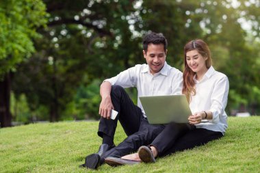 Happy asian couple college working via technology laptop with laughing action in the park, business and lifestyle concept