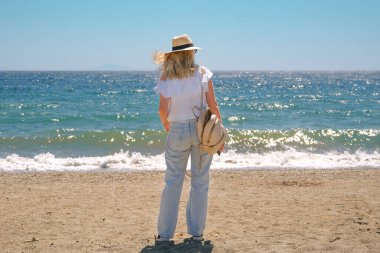 Hasır şapkalı ve kot pantolonlu bir kadının sahile doğru yürüyüşü..