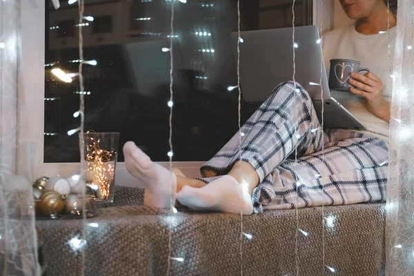 Woman in pajamas sitting on the window sill with her laptop computer. Christmas holidays mood.