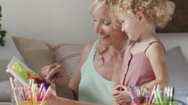 Gülümseyen anne kıvırcık saçlı sarışın kıza gülümsüyor, evde okul öncesi öğrenim aktivitesi, sağlıklı büyüme kavramı, güneşli oturma odasında eğlenceli oyunlar oynayıp yazıyor.