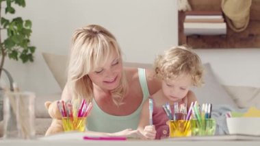 Gülümseyen anne kıvırcık saçlı sarışın kıza gülümsüyor, evde okul öncesi öğrenim aktivitesi, sağlıklı büyüme kavramı, güneşli oturma odasında eğlenceli oyunlar oynayıp yazıyor.