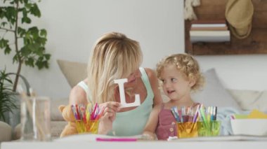 Gülümseyen anne kıvırcık saçlı sarışın kız çocuğuna gülümsüyor, evde okul öncesi öğrenme aktivitesi, sağlıklı büyüme kavramı, güneşli oturma odasında neşeli bir şekilde oynayan aşk kelimesini okuyup yazıyor.