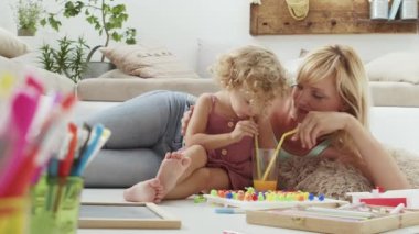 Gülümseyen anne kıvırcık saçlı sarışın kıza gülümsüyor, aynı bardaktan neşeyle içiyor portakal suyunu pipetle bölüyor, evde anaokulu öğrenme aktivitesi, sağlıklı büyüme kavramı.