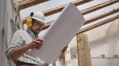 Mimar, miğfer ve kulaklık takıyor. Şablona bakıyor. İnşaat projesinin planını gözden geçiriyor. İnşaat alanının arka planında çatının eski ahşap kirişleri var.