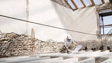 man at work, construction worker wearing helmet and looking at blueprint , check the house project plan, in renovation building site background with the beams at the base of the foundations clipart