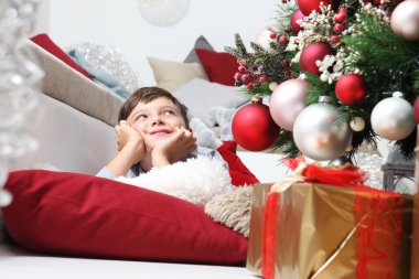 Çocuk ve toplarla süslenmiş Noel ağacı, oturma odasında yerde, paketlenmiş hediyelerin yanında yatıyor.