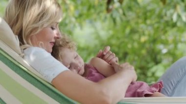 Mutlu anneler günü, gülümseyen anneler günü. Mavi gözlü, kıvırcık sarı saçlı kız çocuğunu kucaklayıp okşar. Hamakta güneşli yeşil bahçede, bahar ve yaz mevsimi konseptinde birlikte uzanır.